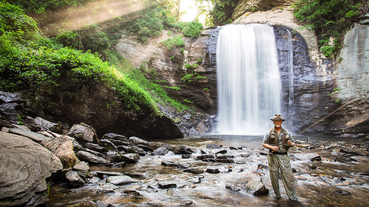 Sam Johnson from Wild Bearings standing ankle deep in rocky moving water with a large waterfall behind him. He is holding a fishing reel and wearing fishing gear